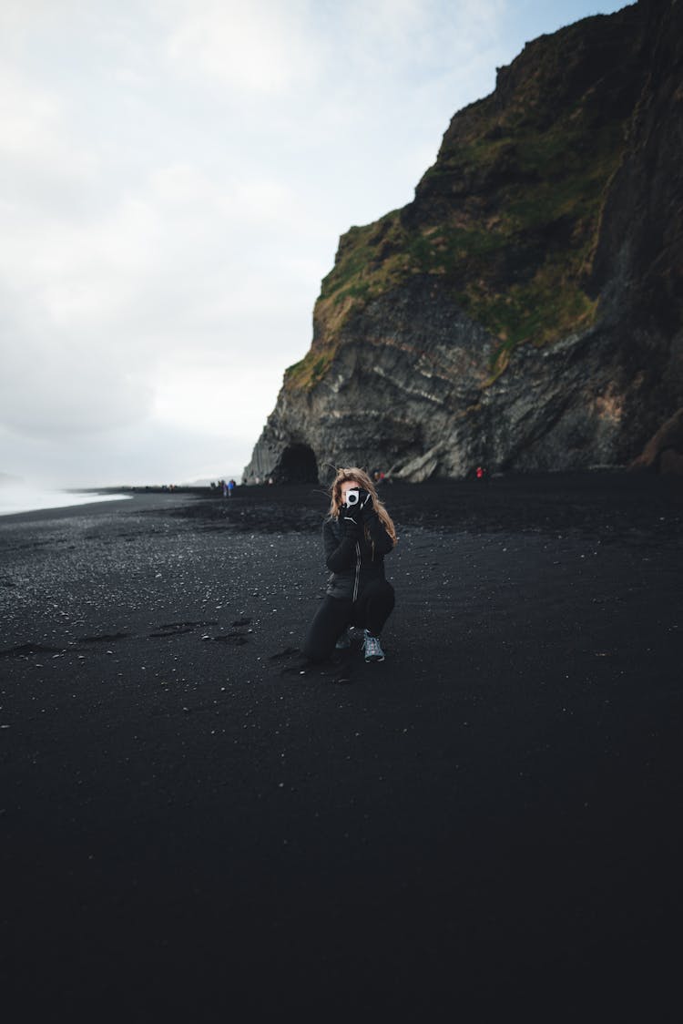 女人跪在岩石峭壁的黑色砾石海滩上拍照